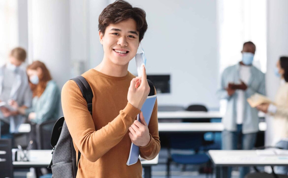 Happy asian guy taking off disposable medical mask at classroom