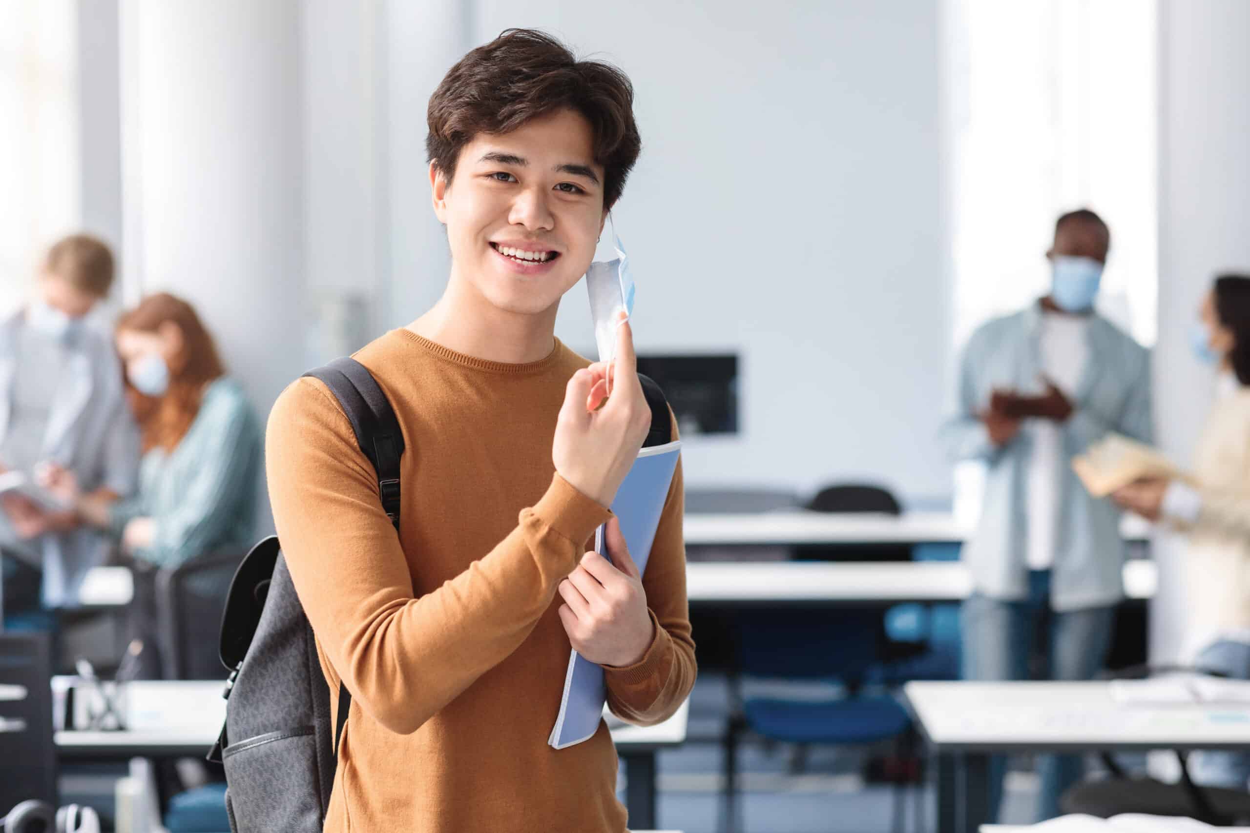 Happy asian guy taking off disposable medical mask at classroom