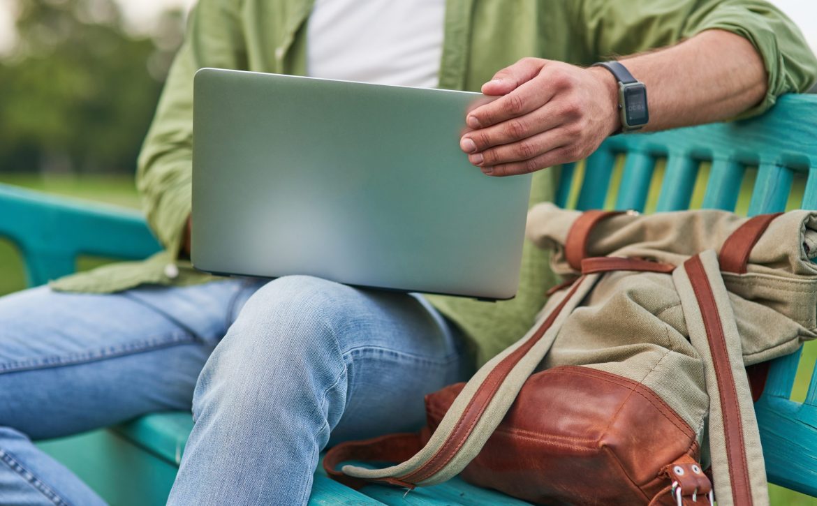 Cropped shot of young man using laptop while working, sitting on the bench outdoors